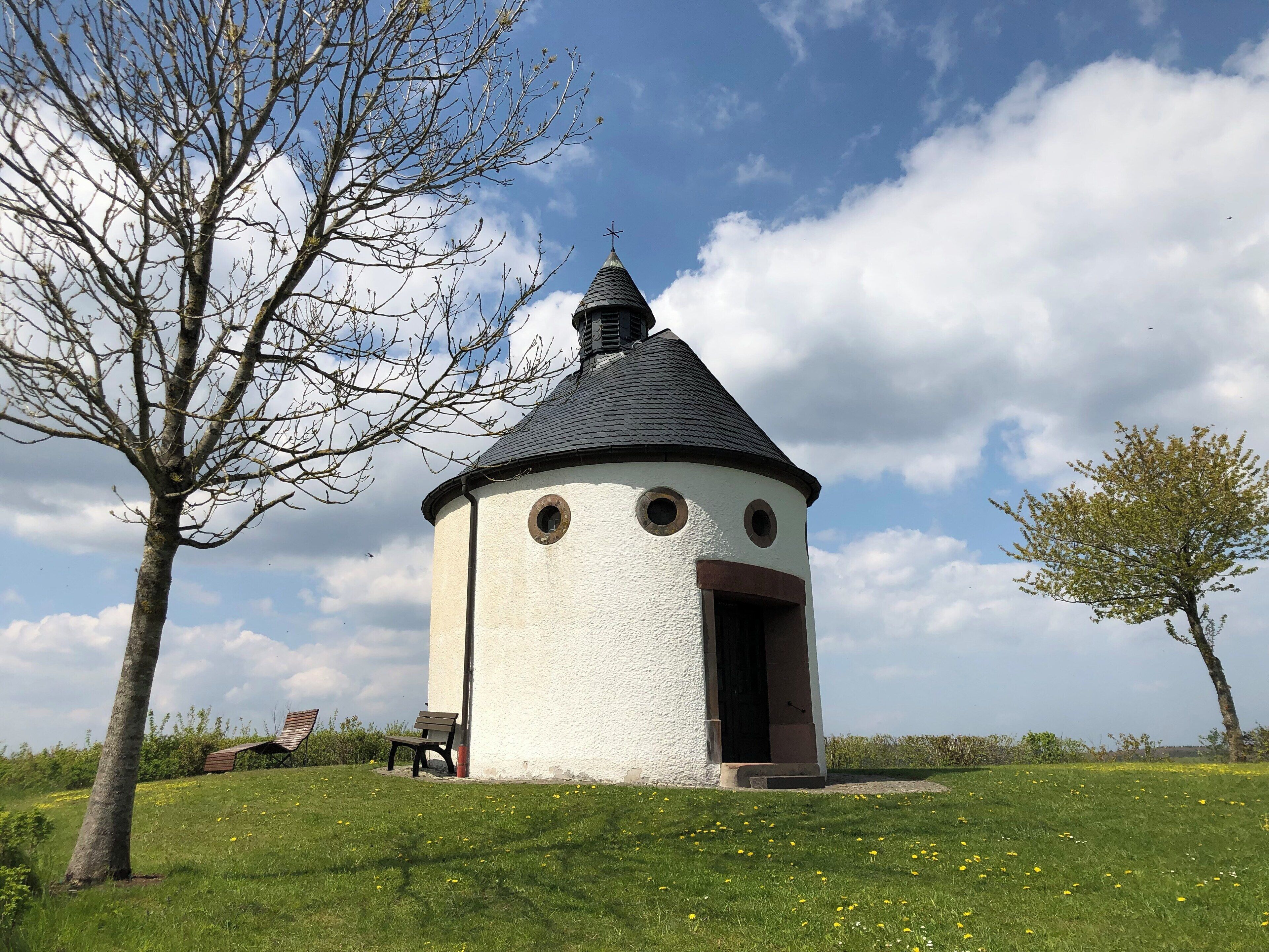 A small white tower with a conical roof and three circular windows, surrounded by a grassy field with scattered yellow flowers and a bench.
