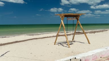Una estructura de madera en la playa con un letrero, sobre arena, con agua turquesa cristalina y un cielo azul con nubes.