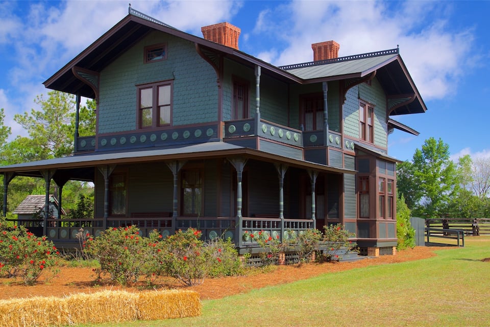Uma casa grande de dois andares com varanda, cercada por um jardim bem cuidado e árvores.