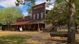 Un edificio de madera rústico con un toldo rojo, identificado como una farmacia, rodeado de árboles y un banco.