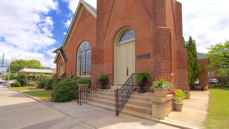 A brick building with a large arched window and a door, surrounded by greenery and a sidewalk.