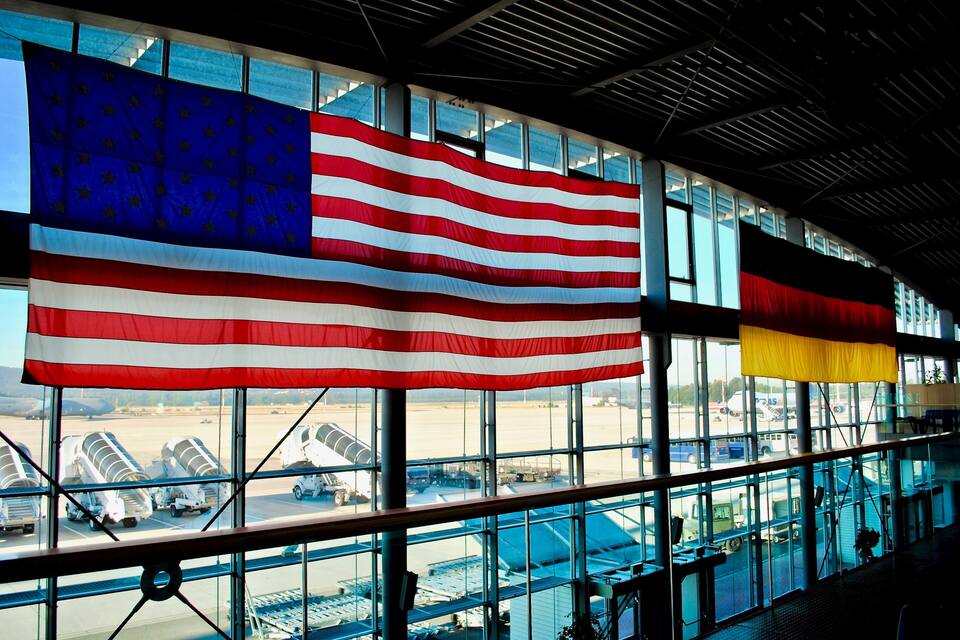 Large American flags and a German flag hanging in a building with large windows overlooking an airport.