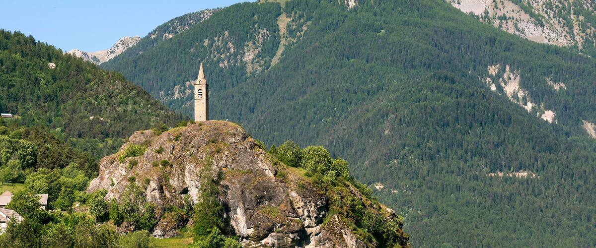 A church with a bell tower on a rocky hill, surrounded by dense forest and mountains.