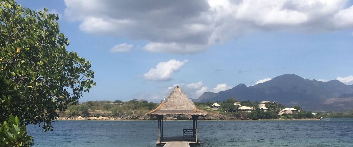 A wooden pier leading to a thatched-roof structure by a calm lake with mountains in the background.