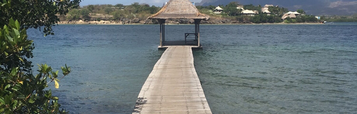 A wooden pier leading to a thatched-roof structure by a calm lake with mountains in the background.