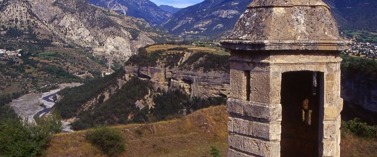 A stone structure with a dome on a hillside overlooking a valley with a river and mountains in the background.
