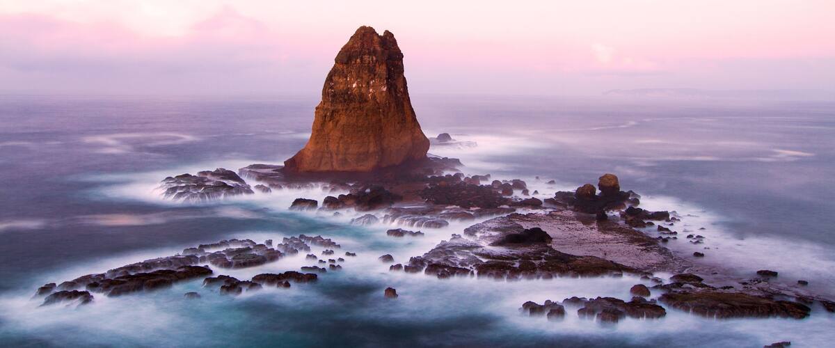 A coastal landscape with a prominent rock formation surrounded by smaller rocks and a misty sea.