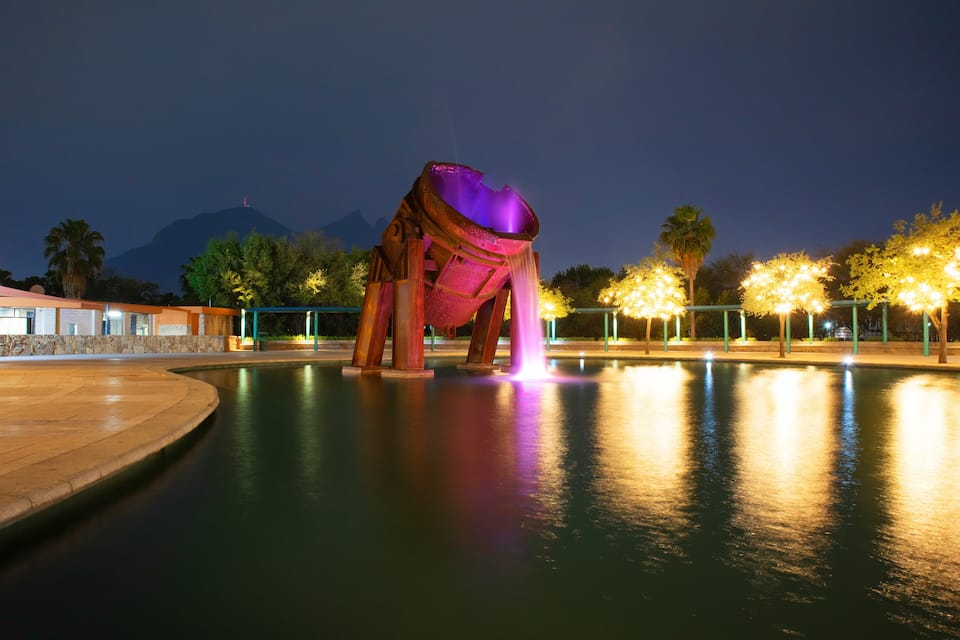 A nighttime scene with a large, illuminated sculpture in a pool, surrounded by trees and a mountain in the background.