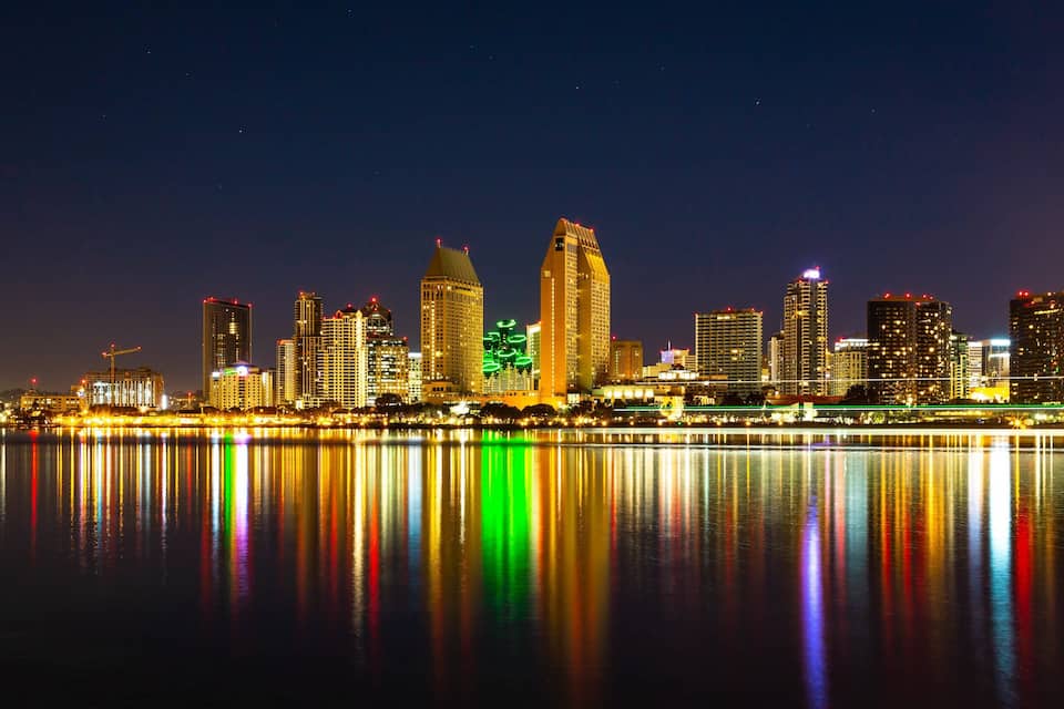 A nighttime city skyline with illuminated buildings reflected in the water.