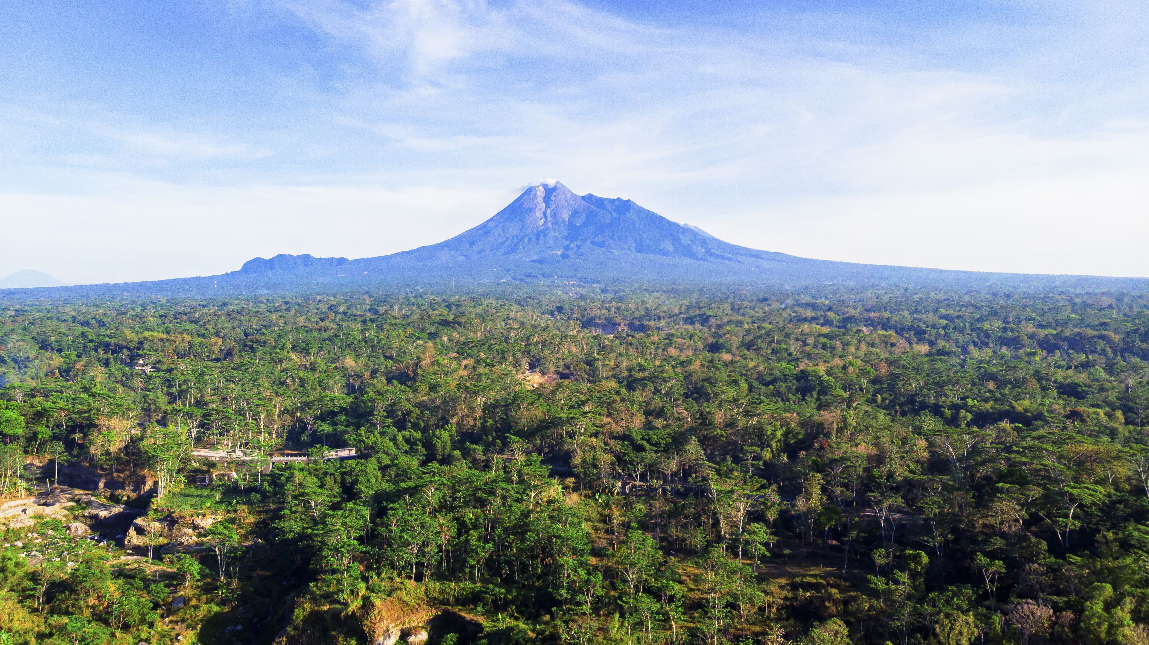 Aerial view of the majestic Merapi Mountain rises distantly over the dense, vibrant green forest, a contrast of cool blues and warm greens, Klaten, Central Java, Indonesia.