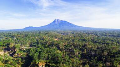 Aerial view of the majestic Merapi Mountain rises distantly over the dense, vibrant green forest, a contrast of cool blues and warm greens, Klaten, Central Java, Indonesia.