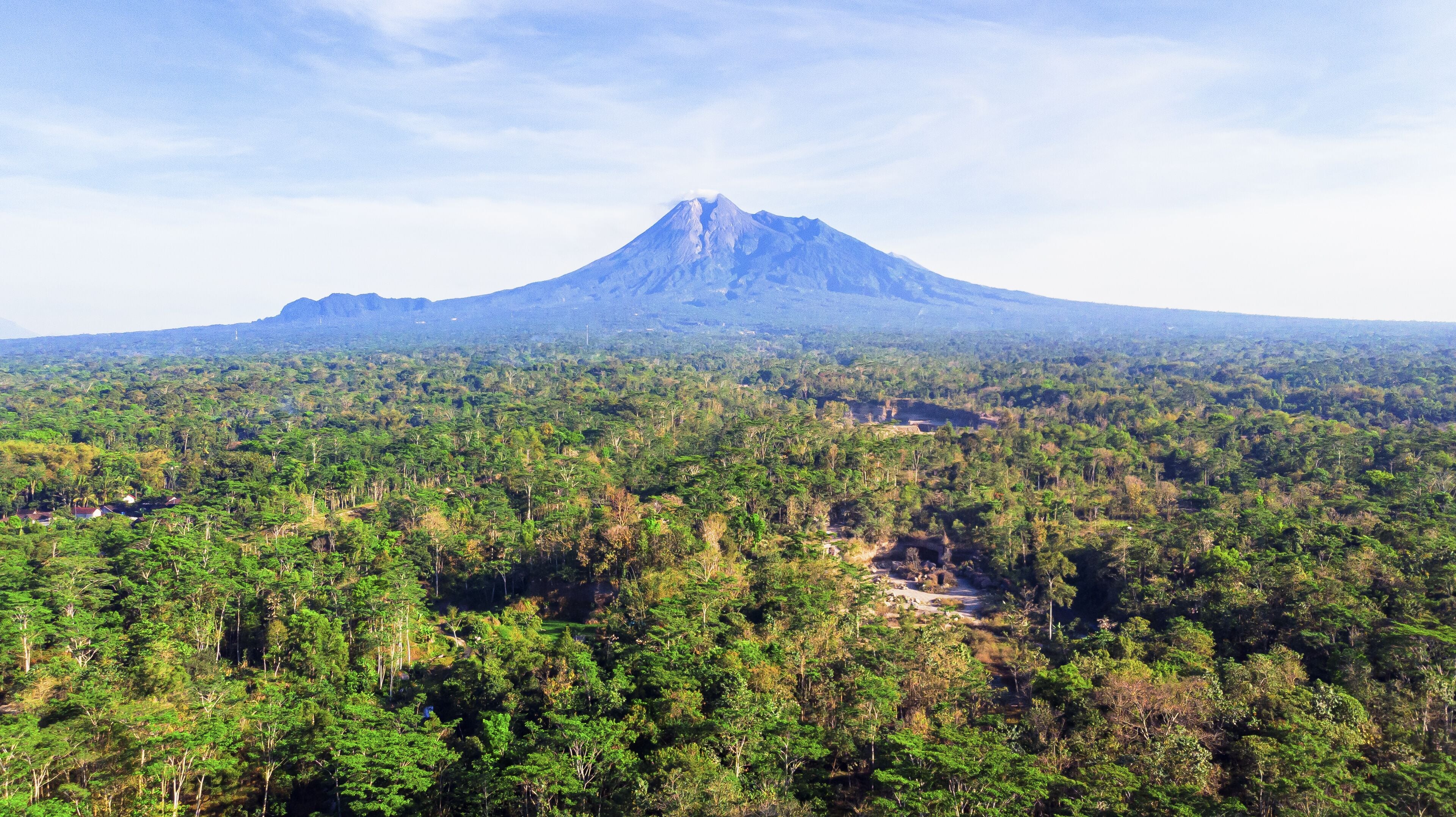 Aerial view of the majestic Mount Merapi rises distantly over a dense tapestry of lush green trees, contrasting against the clear blue sky, Klaten, Central Java, Indonesia.