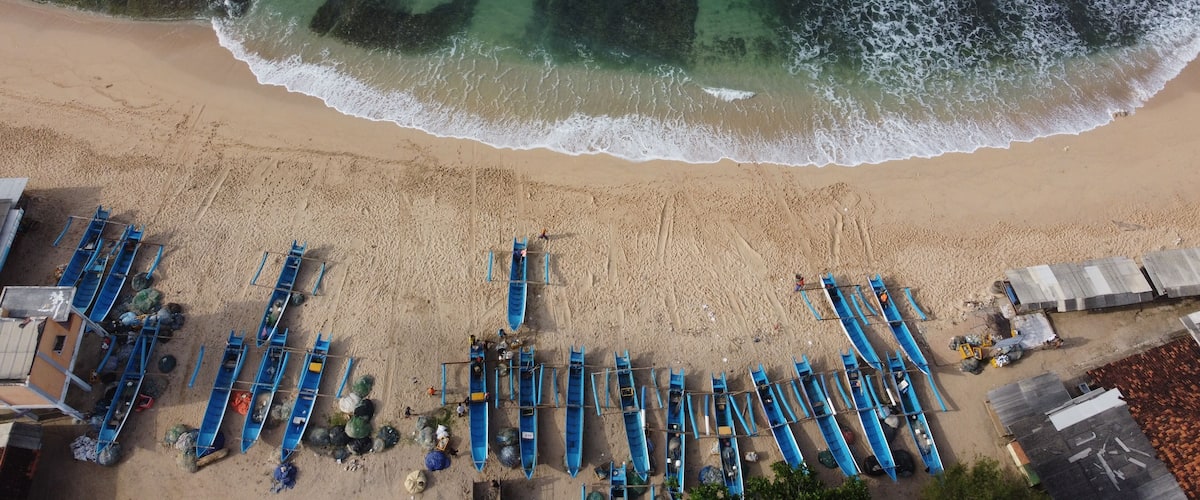 A beach with rows of blue boats lined up on the sand, a sandy shoreline, and the ocean with waves crashing onto the shore.