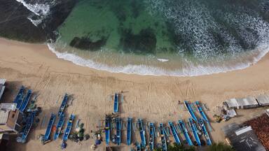 Uma praia com fileiras de barcos azuis na areia, uma linha de costa arenosa e o oceano com ondas quebrando na praia.