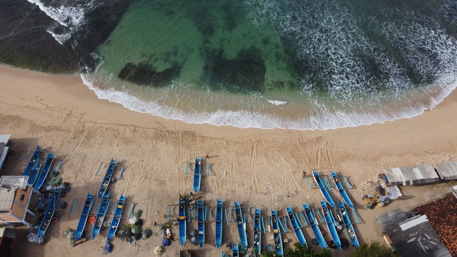 A beach with rows of blue boats lined up on the sand, a sandy shoreline, and the ocean with waves crashing onto the shore.