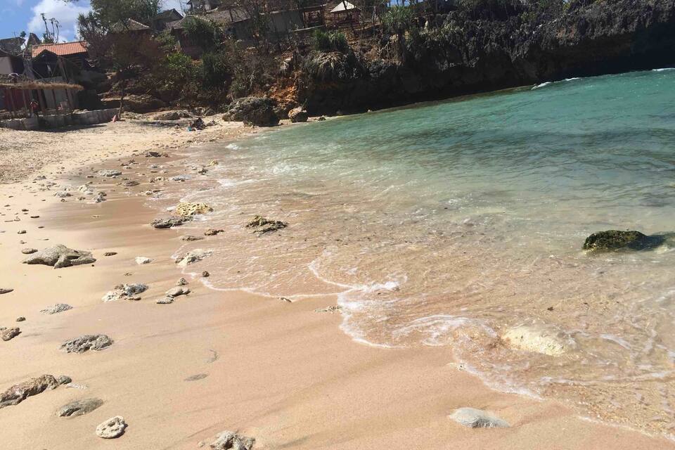 A sandy beach with clear turquoise water, scattered rocks, and houses on a hillside in the background.