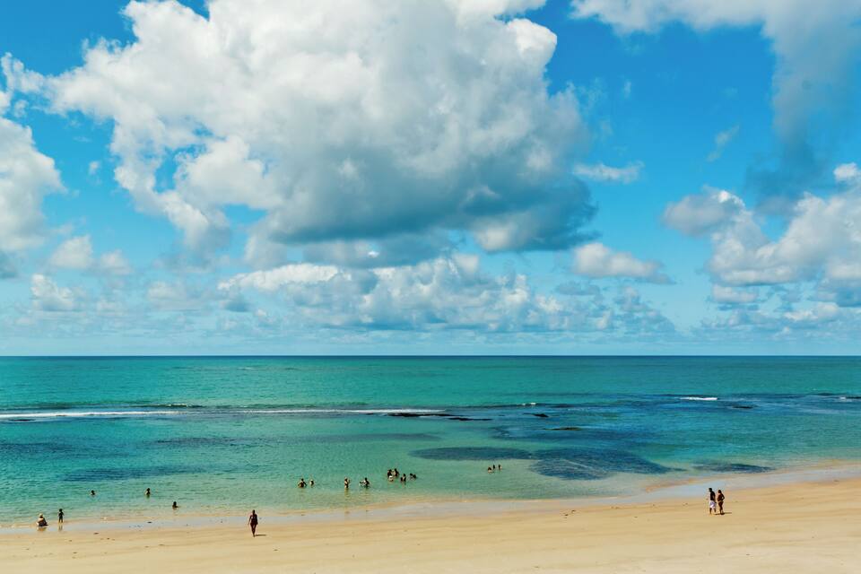 A beach with clear turquoise water, a sandy shore, and a partly cloudy sky.