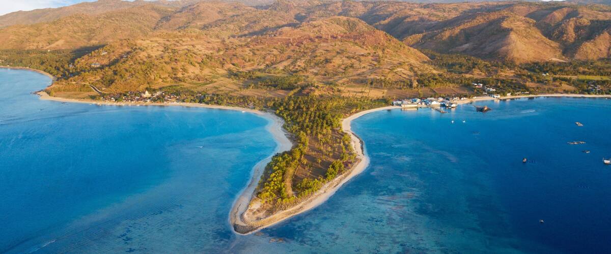 An aerial view of a coastal area with a narrow peninsula surrounded by deep blue waters, a small settlement with houses near the shore, and a backdrop of rolling hills.