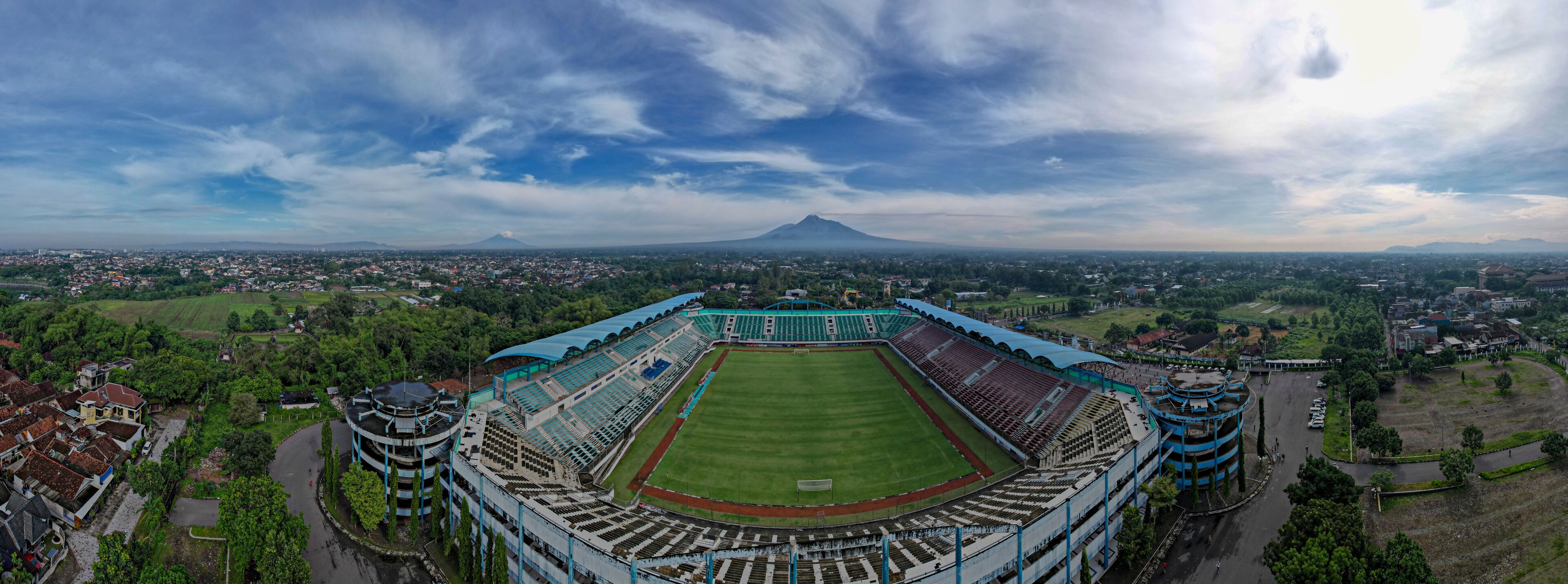 A large stadium with a green field, surrounded by buildings and trees, with a mountain in the background.