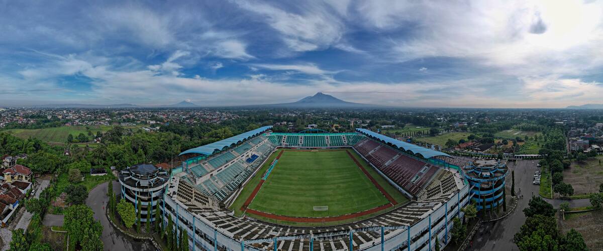 A large stadium with a green field, surrounded by buildings and trees, with a mountain in the background.