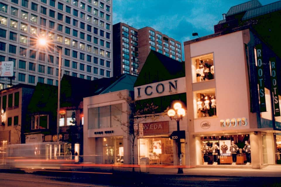Calle urbana al atardecer con escaparates iluminados, incluyendo un llamativo letrero 'ICON' y una tienda de ropa 'ROOTS'.