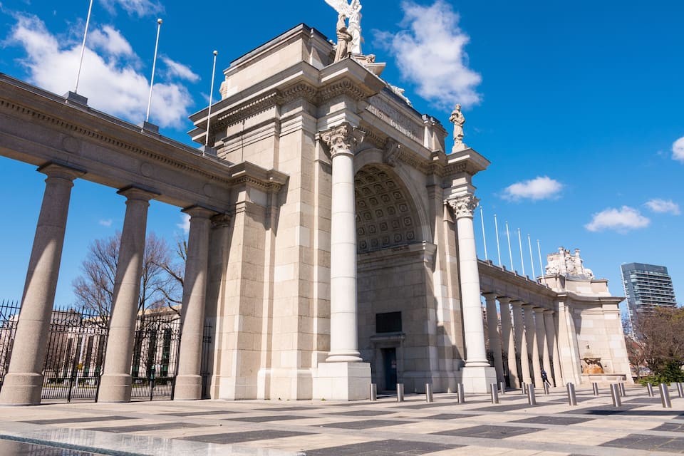 Un imponente edificio neoclásico con amplias columnas y un arco central, flanqueado por estatuas y un alto monumento.