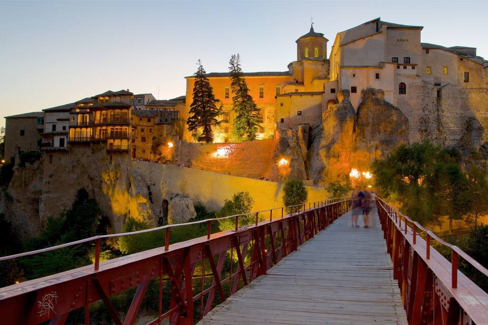 Un ponte di legno conduce a un'antica fortezza in pietra, con edifici illuminati al crepuscolo.