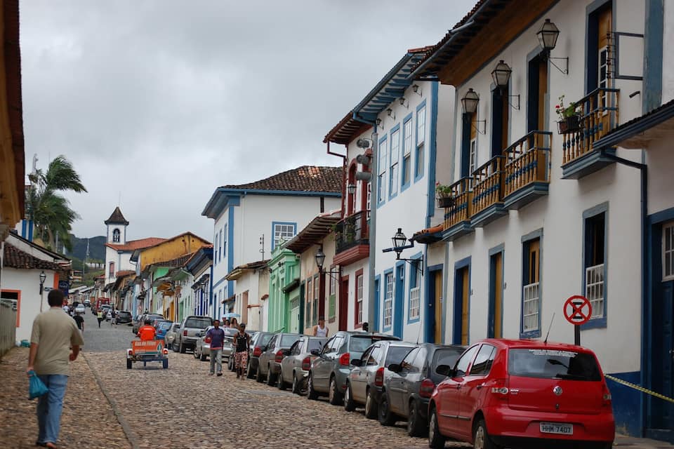 A cobblestone street lined with colorful buildings, parked cars, and a few pedestrians.