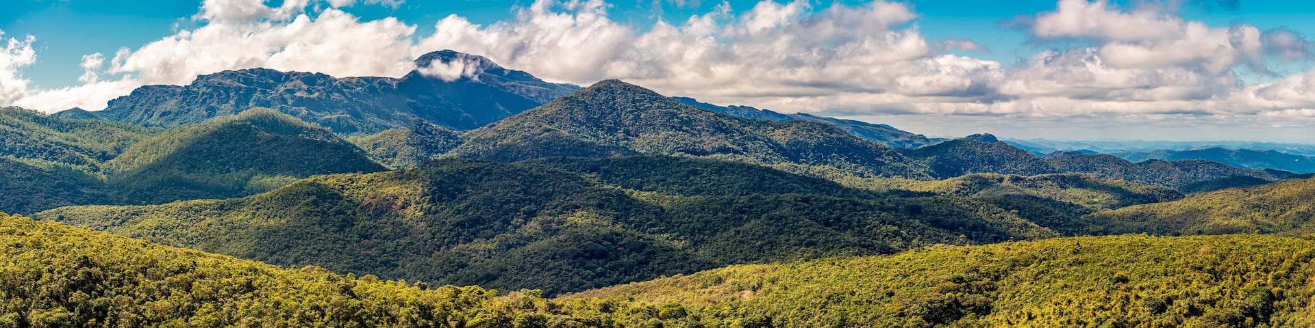A mountainous landscape with dense green forests and a prominent peak under a partly cloudy sky.