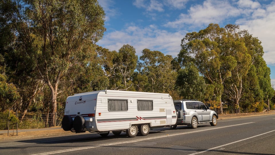 A white RV with a camper trailer attached, driving on a road with trees on both sides.