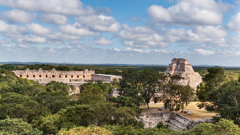 A Mayan ruin with large stone structures and a pyramid, surrounded by dense forest.