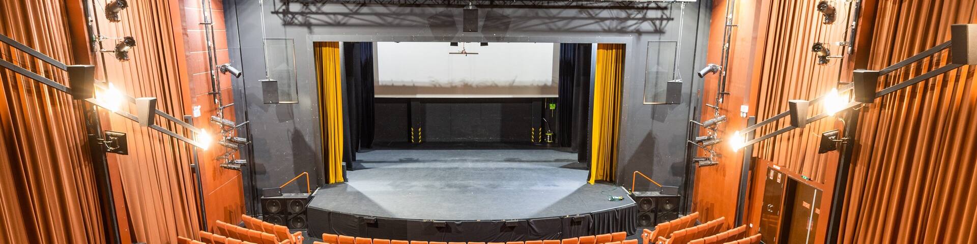 An indoor theater with rows of orange seats, a stage, and a large screen.