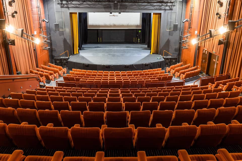 An indoor theater with rows of orange seats, a stage, and a large screen.