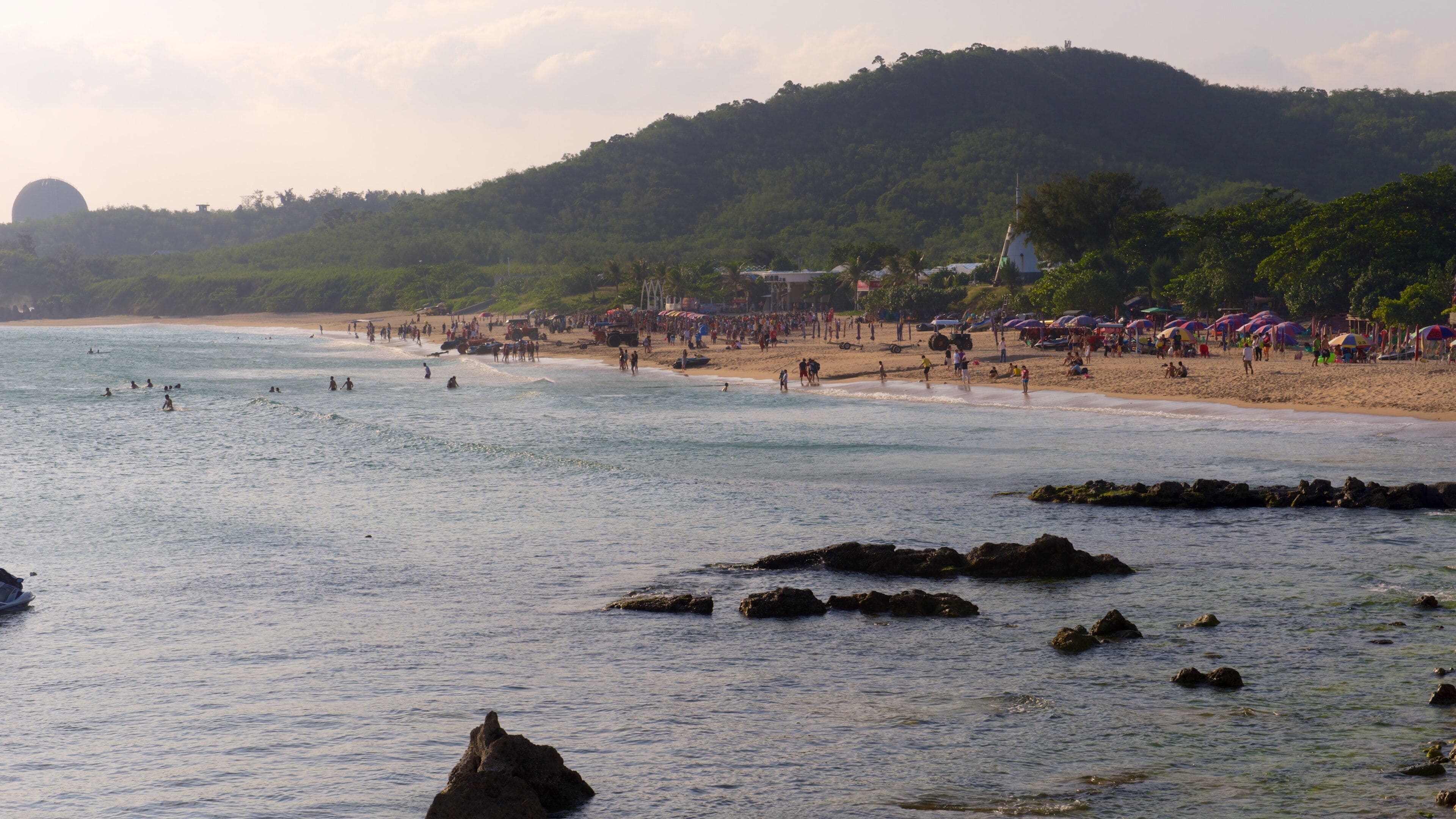 Una playa con gente nadando y tomando el sol, una orilla arenosa y una colina con árboles al fondo.