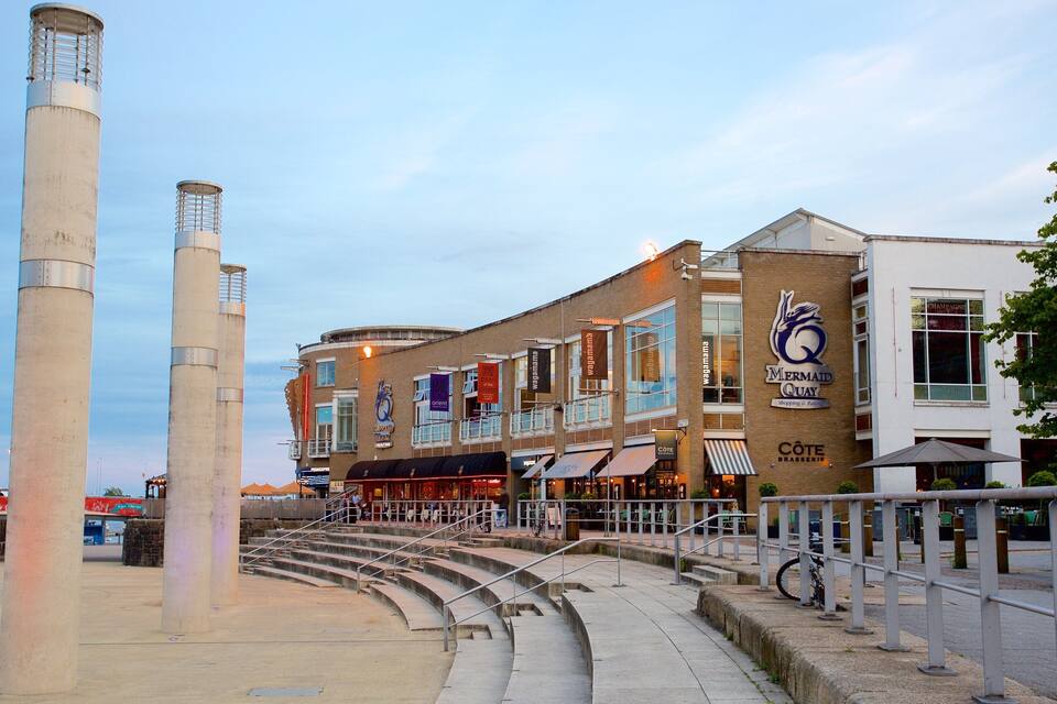 Un paseo marítimo con escalones que conducen a una zona de descanso, una estructura de concreto alta y una hilera de edificios comerciales con diversas tiendas.