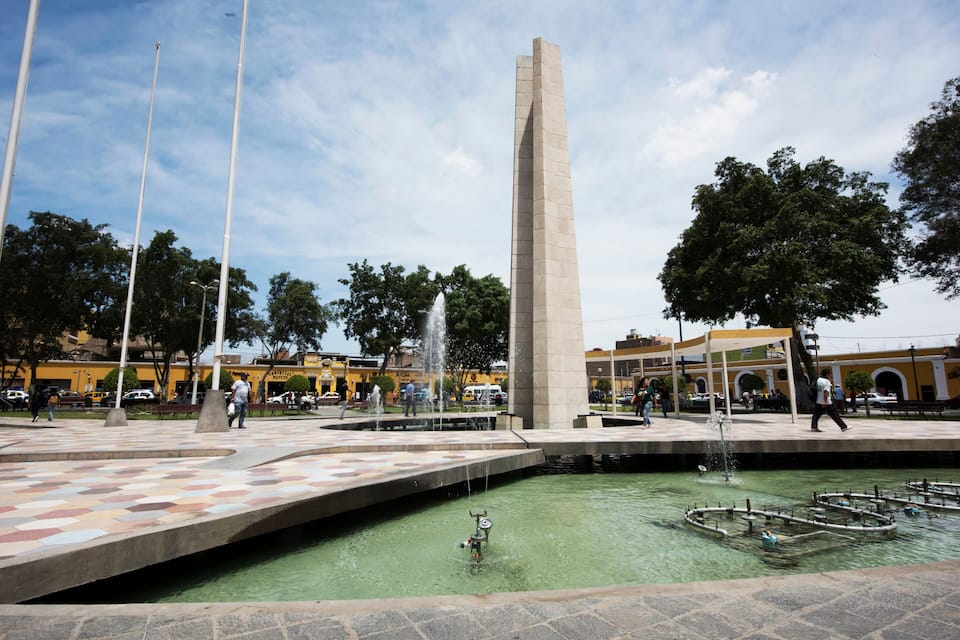 A public square with a tall monument, a fountain, and a patterned tiled area.