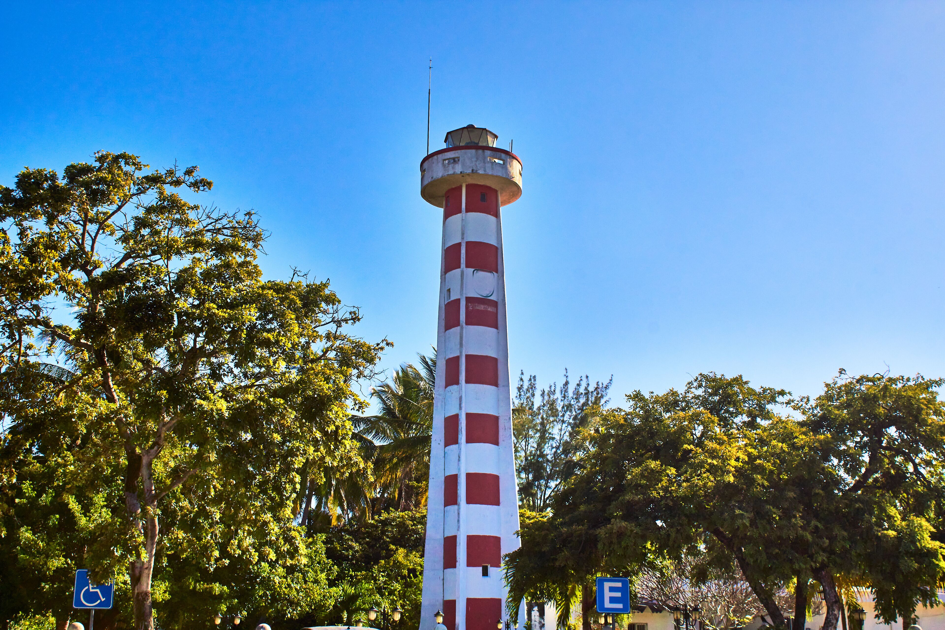 Un alto faro con franjas rojas y blancas, rodeado de frondosos árboles verdes.