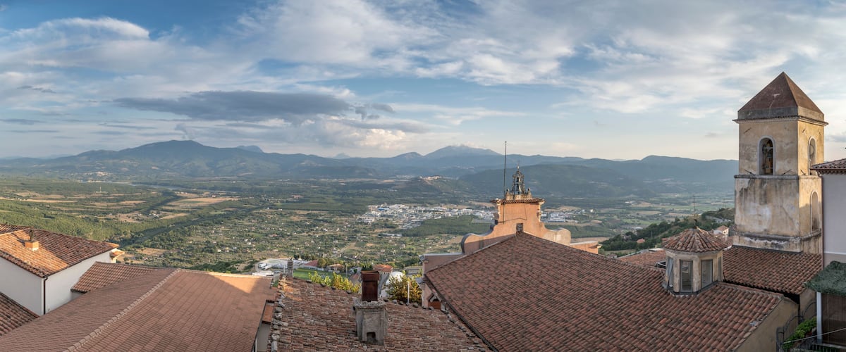 A town with terracotta-roofed buildings, a prominent church tower, and a mountainous landscape in the background.