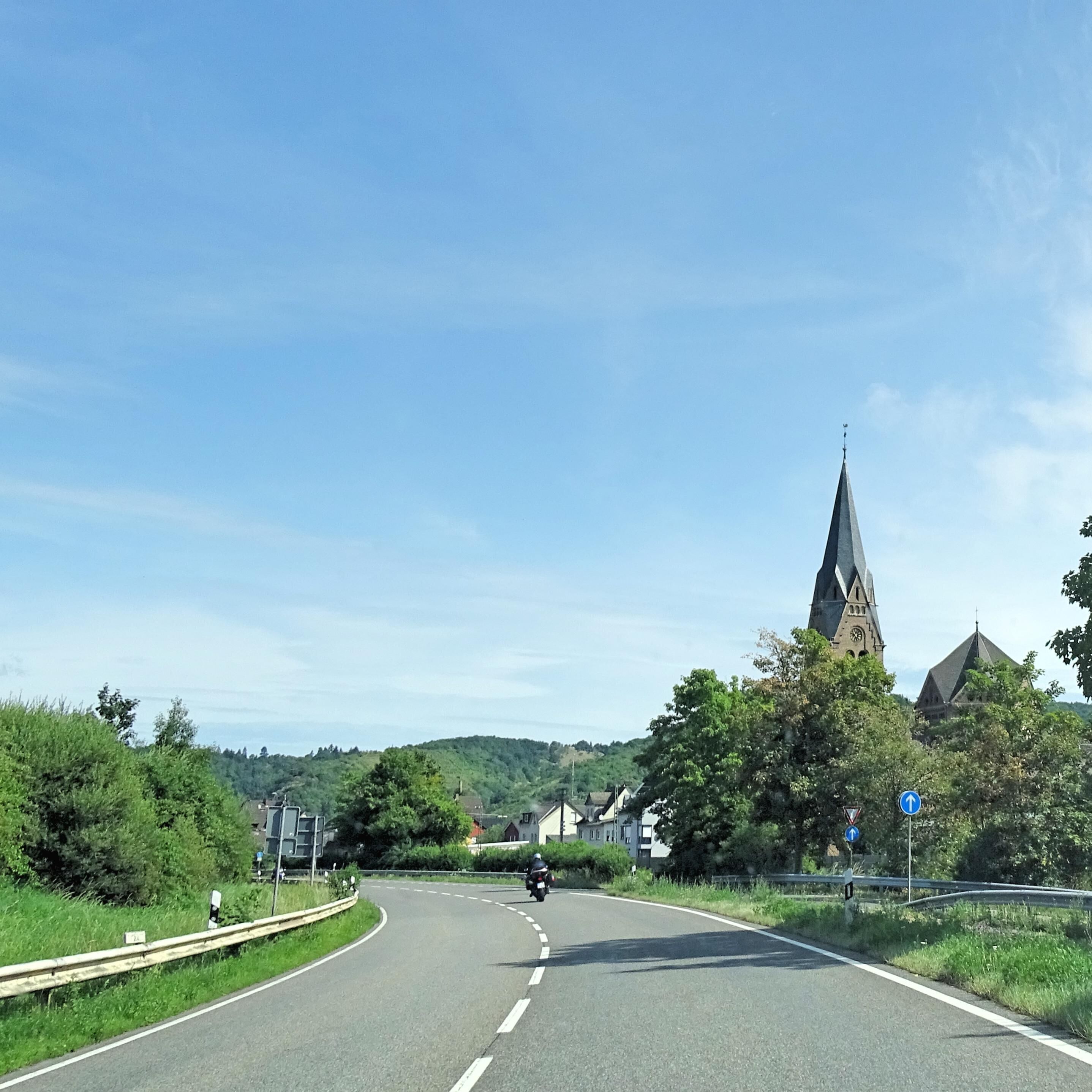 A curved road with a motorcyclist ahead, surrounded by greenery and a church with a tall spire.
