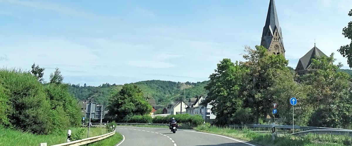 A curved road with a motorcyclist ahead, surrounded by greenery and a church with a tall spire.