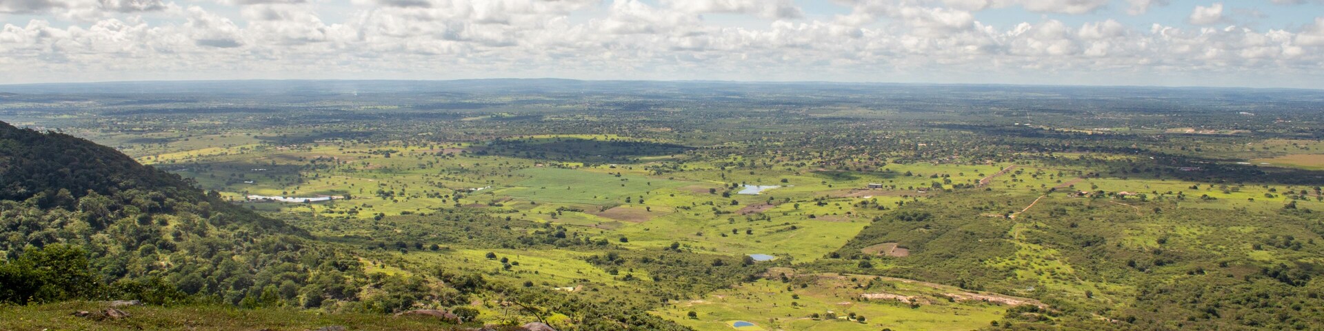 A panoramic view of a lush green valley with scattered trees and a distant mountain range under a partly cloudy sky.