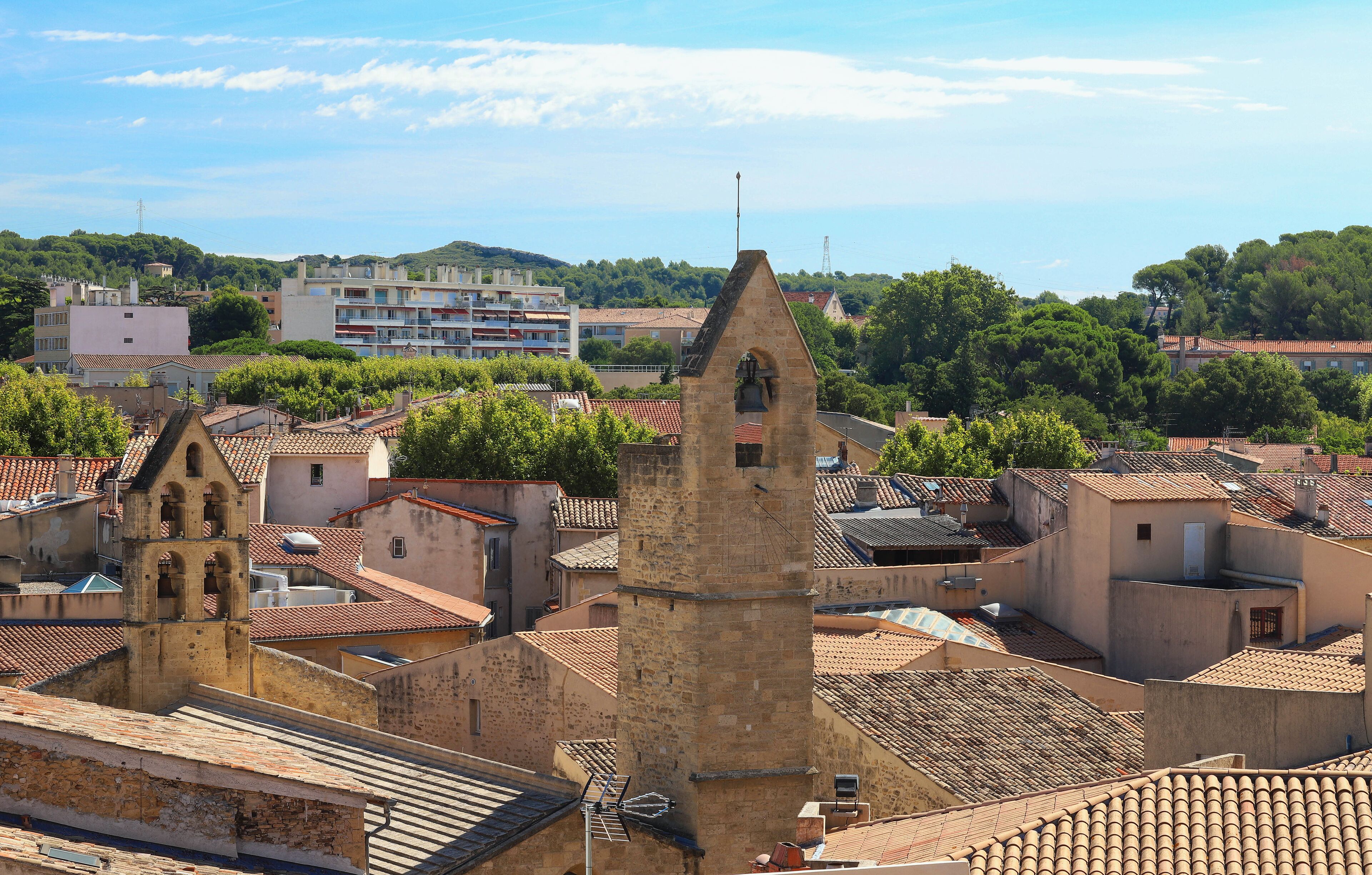 A historic town with a prominent bell tower, terracotta-roofed buildings, and lush green hills in the background.