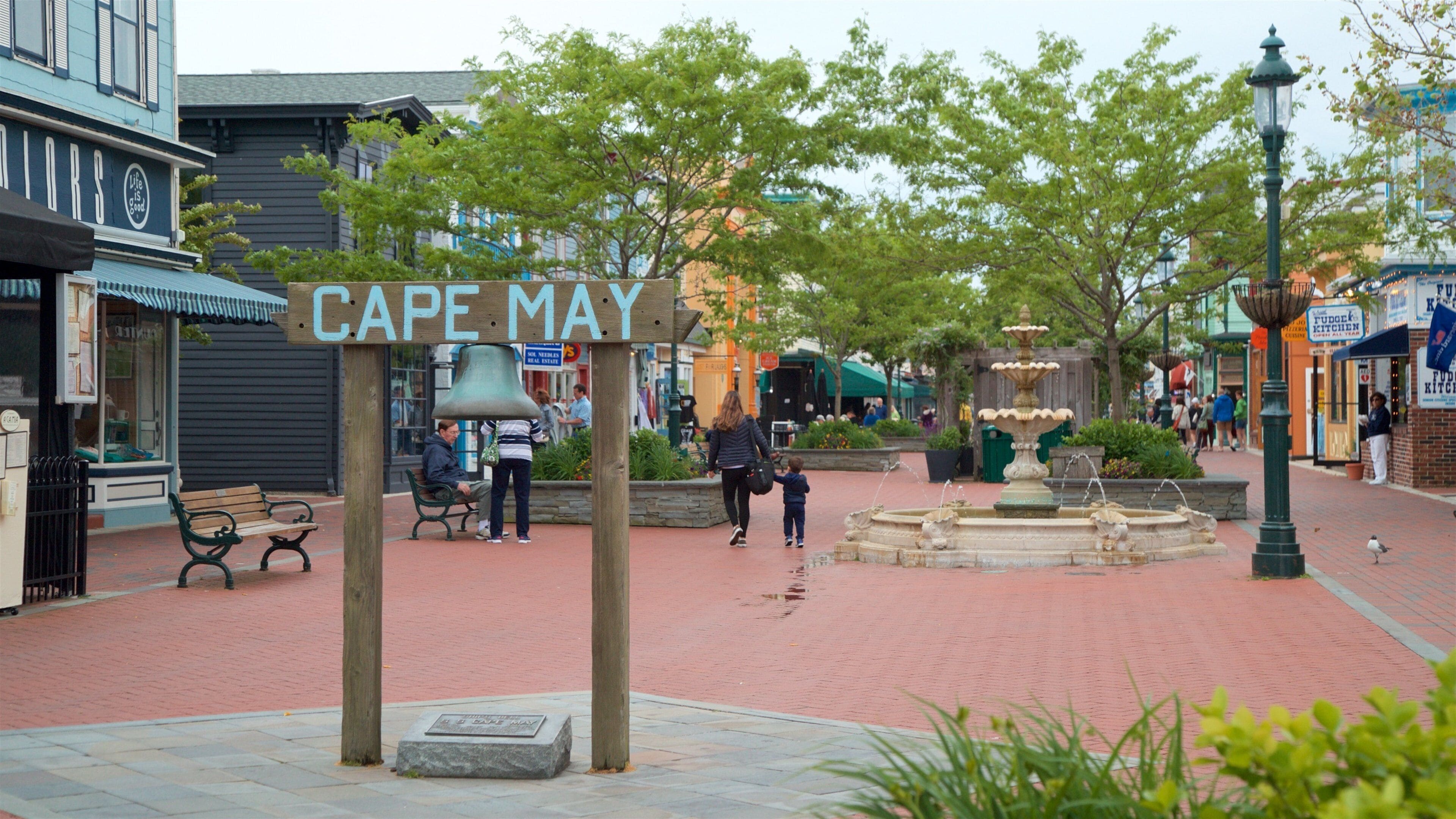 A public square with a sign reading "Cape May," benches, a fountain, and surrounding shops.