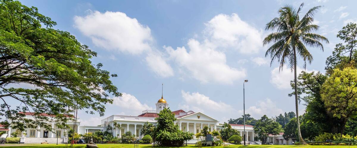 A large white building with a golden dome, surrounded by lush greenery and a calm pond with lily pads.