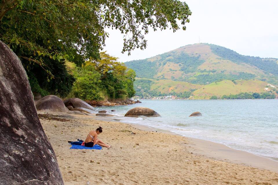 Une plage où une personne prend un bain de soleil, avec de gros rochers et des collines en arrière-plan.