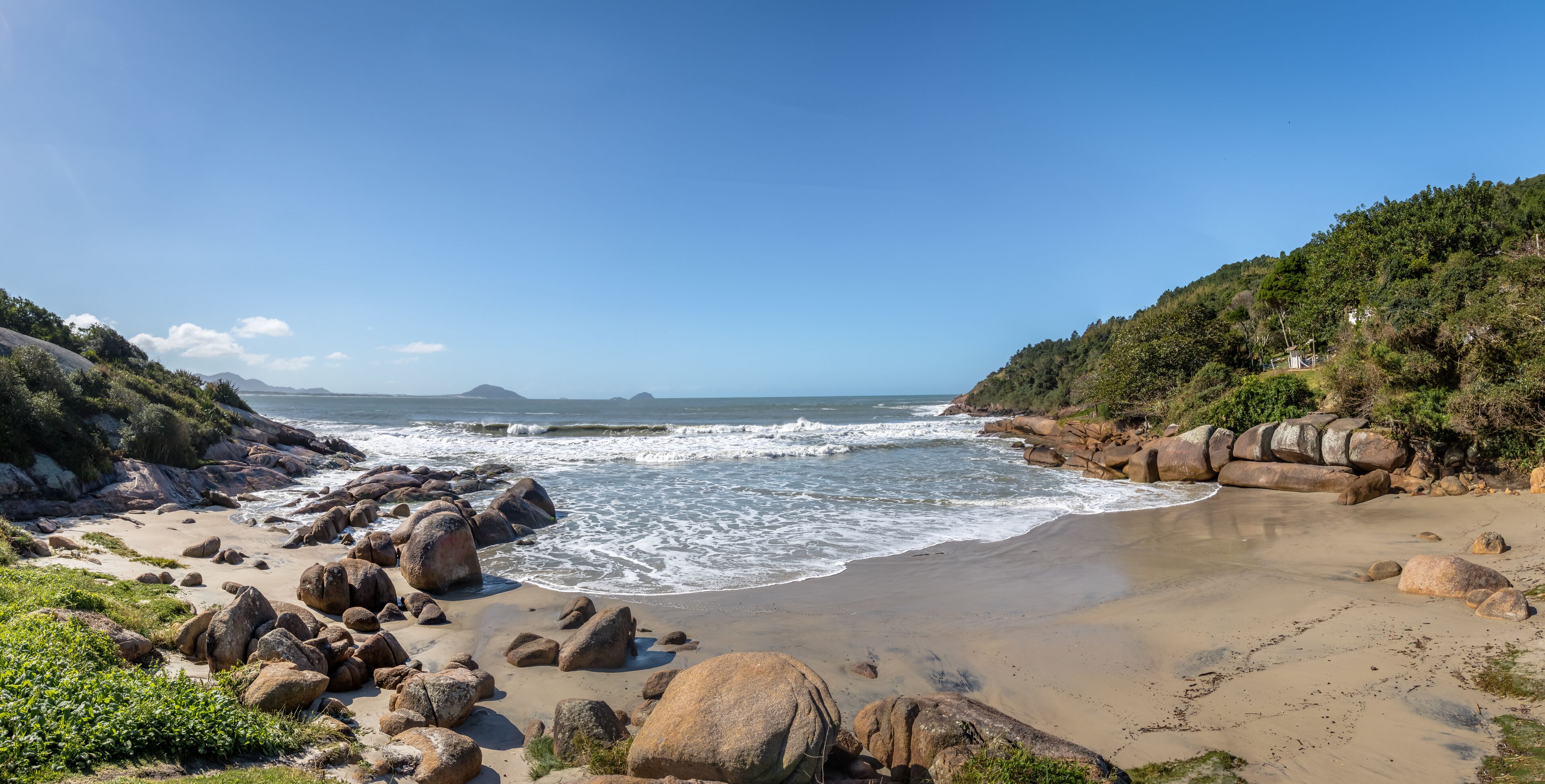 A rocky beach with waves crashing onto the shore, surrounded by hills and a clear blue sky.