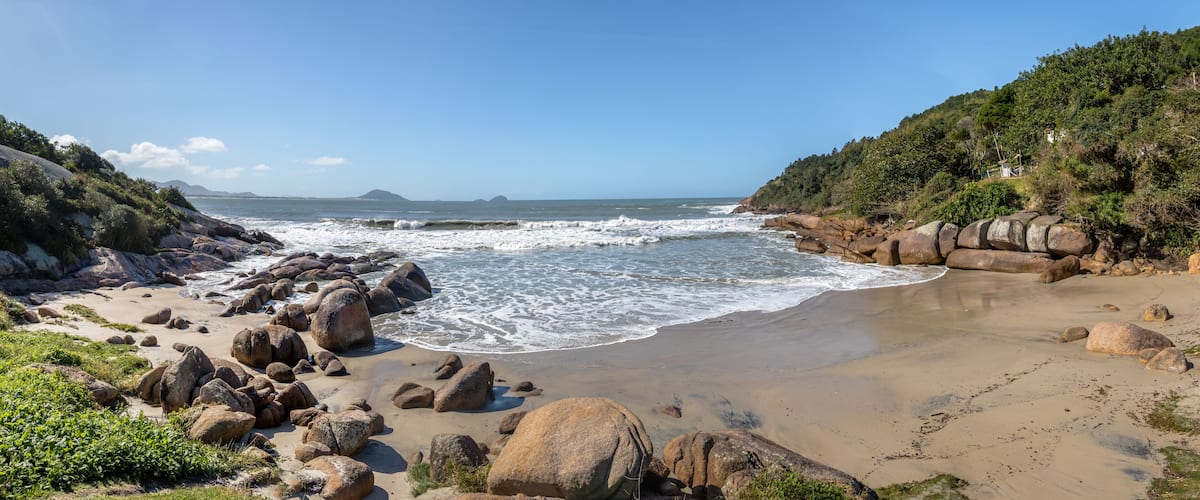 A rocky beach with waves crashing onto the shore, surrounded by hills and a clear blue sky.