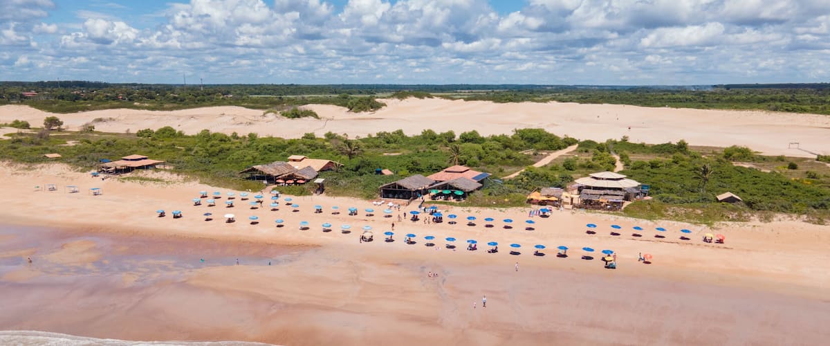 Ein Strand mit vielen Sonnenschirmen, Menschen, üppiger Vegetation und Gebäuden.