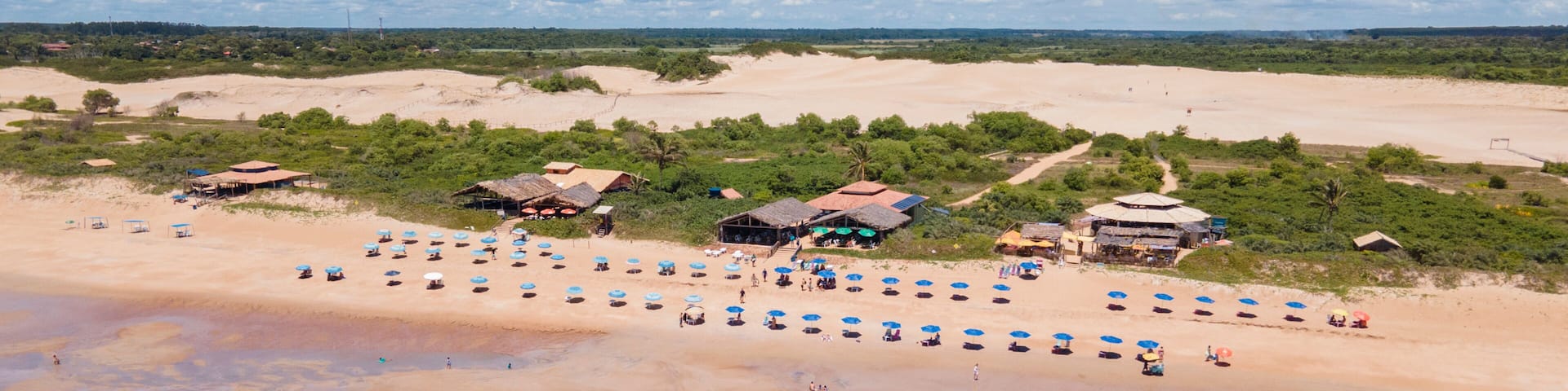 A beach with numerous umbrellas and people, surrounded by greenery and buildings.