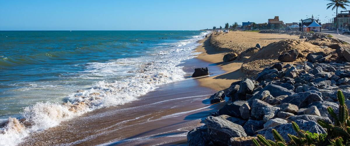 A beach with waves, rocks, and buildings.